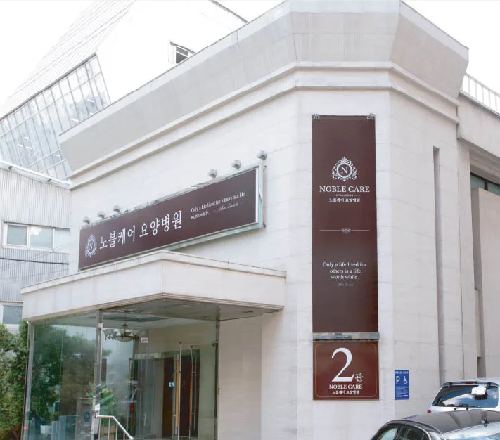 노블케어요양병원 2관 외관. 흰 석재 벽면에 세로 배너와 2관 표시가 붙어 있다