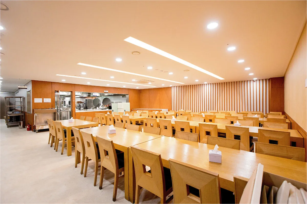 식당. 긴 나무 식탁과 의자가 줄지어 있고 배식 창구가 보인다
