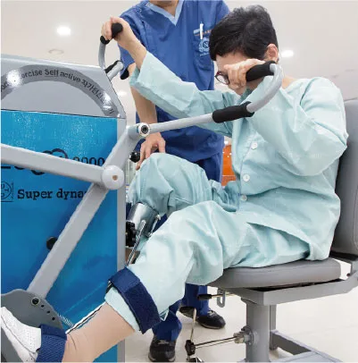 환자가 하지 운동기구에 앉아 다리 운동을 하고 치료사가 옆에서 돕고 있다